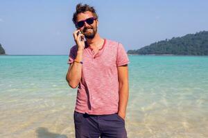glücklich Reisen Mann mit Bart reden durch Handy, Mobiltelefon Telefon und bleiben im Blau Meer Wasser auf seine tropisch Feiertage. halten Telefon im Hände. voll Länge. foto