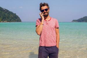 stilvoll Mann mit Bart mit Smartphone auf seine tropisch Urlaube. sonnig Strand Hintergrund. Reisender Hipster. Urlaub arbeiten. Senden Botschaft. foto