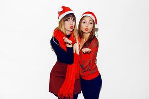 Neu Jahr Party. zwei schön Mädchen im komisch Maskerade Santa Hüte senden Kuss . Innen- Vorrat Bild von Beste freunde posieren auf Weiß Hintergrund. isolieren. foto