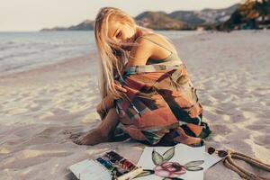 Sommer- Lebensstil Bild von blond Frau Zeichnung Aquarell Blume durch Bürste. Künstler Sitzung auf das Strand. Bohemien Outfit. foto