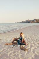 Sommer- Lebensstil Bild von blond Frau Zeichnung Aquarell Blume durch Bürste. Künstler Sitzung auf das Strand. Bohemien Outfit. foto