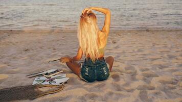 Sommer- Lebensstil Bild von blond Frau Zeichnung Aquarell Blume durch Bürste. Künstler Sitzung auf das Strand. Bohemien Outfit. foto