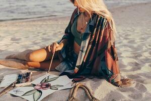 Sommer- Lebensstil Bild von blond Frau Zeichnung Aquarell Blume durch Bürste. Künstler Sitzung auf das Strand. Bohemien Outfit. foto