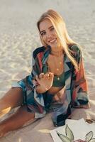 Sommer- Lebensstil Bild von blond Frau Zeichnung Aquarell Blume durch Bürste. Künstler Sitzung auf das Strand. Bohemien Outfit. foto