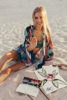 Sommer- Lebensstil Bild von blond Frau Zeichnung Aquarell Blume durch Bürste. Künstler Sitzung auf das Strand. Bohemien Outfit. foto