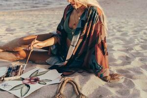 Sommer- Lebensstil Bild von blond Frau Zeichnung Aquarell Blume durch Bürste. Künstler Sitzung auf das Strand. Bohemien Outfit. foto