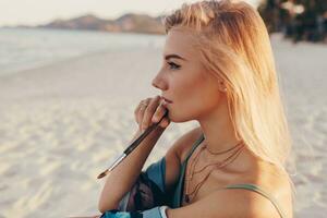 Sommer- Lebensstil Bild von blond Frau Zeichnung Aquarell Blume durch Bürste. Künstler Sitzung auf das Strand. Bohemien Outfit. foto