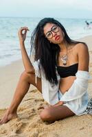 schließen oben Lebensstil Porträt von stilvoll asiatisch Frau mit rot Lippen. sie ist Sitzung auf sonnig tropisch Strand. Boho Accessoires.Schmuck ,Armband und Halskette. foto