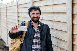 heiter Hipster Bart Mann lächelnd, Herstellung selbst Porträt durch Handy, Mobiltelefon Telefon auf Holz Mauer Hintergrund. tragen stilvoll schwarz Hut, Leder Jacke und Rucksack. foto