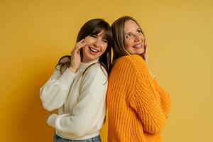 schließen oben Porträt von zwei ziemlich Frau im gemütlich Pullover posieren Über Gelb Hintergrund. Herbst und Winter Mode Tendenzen. foto