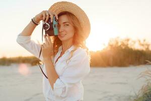 Reisen und Ferien Konzept. glücklich weiblich Gehen auf Abend Strand und halten retro Kamera. tragen Stroh Hut und sexy Jeans kurze Hose. warm Sonnenuntergang Farben . foto