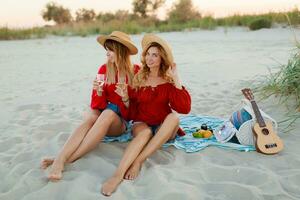 zwei ziemlich Frauen im rot Sommer- Outfit abd Stroh Hüte genießen Picknick auf das Strand. Sommer- Stimmung. foto