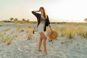 ziemlich Brünette Frau im stilvoll Boho Sommer- tragen Gehen auf das Strand. warm Sonnenuntergang Farben.Stroh Hut. foto