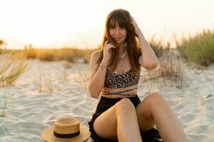 sexy Frau im stilvoll Sommer- Boho Outfit Sitzung auf Sand und genießen Ferien auf das Strand. Sonnenuntergang Farben. foto