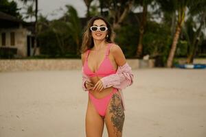 schön stilvoll Frau im modisch Rosa Badebekleidung posieren auf das Strand. Sommer- Urlaub, tropisch Insel, Palmen. tragen beiläufig Sonnenbrille. foto