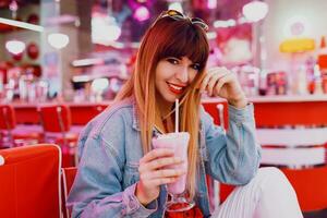 modisch Mädchen im Jeans Jacke Sitzung im t retro amerikanisch Cafe. schön lächelnd Frau genießen Süss Milch Shake . tragen Weiß Turnschuhe. foto