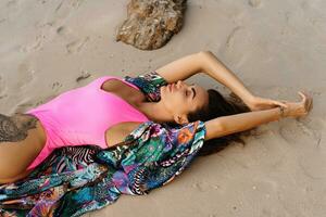 sinnlich schlank Frau mit perfekt Körper Lügen auf Sand auf tropisch Insel. tragen stilvoll Rosa Badebekleidung und Tunika mit tropisch drucken. foto