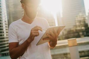 draussen Porträt von gutaussehend Mann im Weiß T-Shirt mit Tablette , suchen im Internet, Stehen Über moden Stadt Hintergrund mit Wolkenkratzer. foto