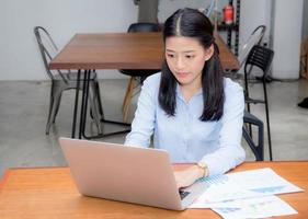 junge asiatische Frau, die mit Diagrammstatistikpapier auf Laptop arbeitet.
