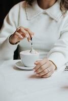 ein Nahansicht Frau Hand rührt sich das Kaffee mit ein Löffel. Cappuccino mit Schokolade auf ein Weiß Tisch. heiß Kaffee trinken Konzept. Restaurant foto