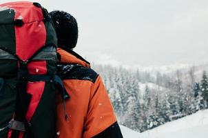 Rückansicht des touristischen Mannes, der die Bergwaldlandschaft an einem verschneiten Wintertag genießt. orangefarbenes Kleidungsstück, roter Rucksack. Wandern Reisen extremes Lifestyle-Konzept foto