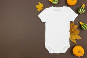 Weiß Baby Mädchen oder Junge Bodysuit eben legen Attrappe, Lehrmodell, Simulation mit Kürbisse und gefallen Blätter auf dunkel Hintergrund. Design Strampler Vorlage, drucken Präsentation spotten hoch. oben Sicht. foto