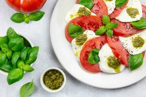 traditionell Italienisch Caprese Salat mit geschnitten Tomaten, Mozzarella, Basilikum, Pesto Soße und würzen auf ein Licht grau Beton Hintergrund. oben Sicht. schließen hoch. foto