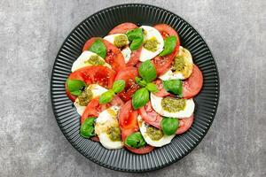 traditionell Italienisch Caprese Salat mit geschnitten Tomaten, Mozzarella, Basilikum, Pesto Soße und würzen auf ein dunkel grau Beton Hintergrund. oben Sicht. foto