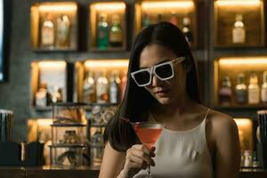 asiatisch Frau Trinken ein Cocktail beim ein Bar während tragen Sonnenbrille. foto