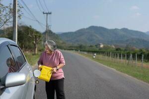 das Auto lief aus von Gas und ins Stocken geraten neben das Straße im Vororte und ein Alten asiatisch Frau benutzt ein Gallone von schonen Gas zu Treibstoff das Wagen. ein Frau bereitet vor ein Gallone von schonen Gas zu Treibstoff Vor reisen. foto