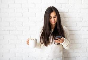 Frau, die eine Tasse Marshmallow-Kakao hält und ihr Handy benutzt foto