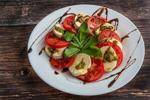 Weiß Teller von gesund klassisch köstlich Caprese Salat mit Tomaten und Mozzarella Käse mit Basilikum Blätter und Streifen von Soja Soße auf das Holz Hintergrund. Italienisch Konzept. foto