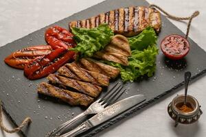 gegrillt Truthahn Brust Steaks mit gebacken Paprika, Salat und Tomate auf ein schwarz Tafel mit Messer und Gabel. Nächster zu das Pfeffer Shaker mit rot Pfeffer. horizontal Orientierung, Nahansicht foto