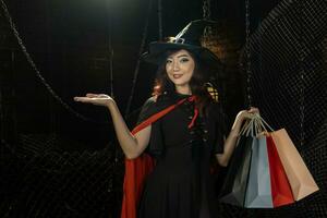 asiatisch Mädchen im Hexe Kostüm Einkaufen Tasche zum Verkauf Beförderung Halloween Party Trick oder behandeln Konzept mit dunkel schwarz Hintergrund foto