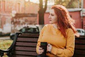 rothaarige frau im gelben sweatshirt sitzt auf der parkbank und trinkt kaffee. sie hält einen wiederverwendbaren becher in ihren händen. Porträt an einem sonnigen Herbsttag. foto