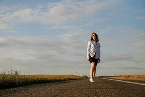Frau in weißem Hemd geht die Straße entlang zwischen den Feldern