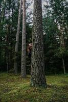 eine junge Frau in einem Nadelwald schaut hinter einem Baum hervor foto