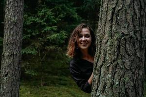 eine junge Frau in einem Nadelwald schaut hinter einem Baum hervor foto