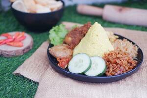 indonesisch Stil Gelb Reis mit Hähnchen und Seite Geschirr im schwarz Teller auf Grün Gras Hintergrund foto