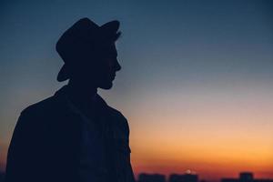 Silhouette eines Männerporträts mit Hut, der den Sonnenuntergang über der Stadt beobachtet