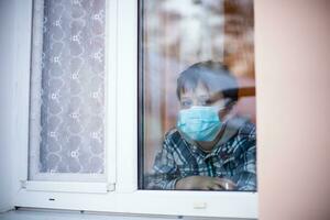 Kind im schützend medizinisch Maske sieht aus aus von das Fenster in Straße. bleibe heim. Isolierung während Coronavirus Epidemie. Schule Quarantäne. foto