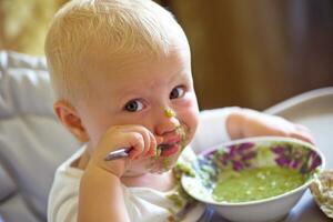 ein wenig Mädchen ist Essen Brokkoli Suppe während Sitzung im ein hoch Stuhl im das Küche foto