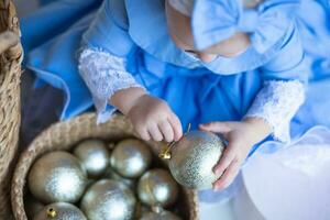 wenig Mädchen im Clever Blau Kleid nimmt aus Weihnachten Ball von Korb. Korb mit Neu Jahre Spielzeuge. vorbereiten zum Neu Jahr und Weihnachten. Kind dekoriert ein Weihnachten Baum. foto