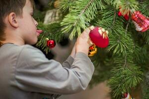 Junge haftet ein rot Ball auf ein Ast von ein Weihnachten Baum beim heim. vorbereiten zum Weihnachten beim heim. echt Personen. dekorieren Weihnachten Baum mit ein Vielfalt von Spielzeuge foto