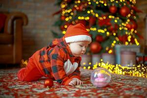 wenig glücklich Mädchen von 6 Monate kriecht in der Nähe von ein Neu Jahr Baum auf Weihnachten Vorabend. Kind im Santa claus Kostüm in der Nähe von leuchtend Girlanden. glücklich Neu Jahr 2020 Konzept foto