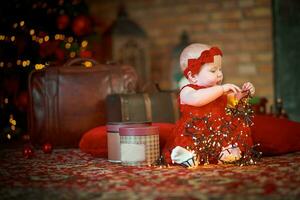 wenig Mädchen im rot Kleid gegen Hintergrund von Weihnachten Baum hält Weihnachten Girlande im ihr Hände. Baby 6 Monat alt feiert Weihnachten. foto