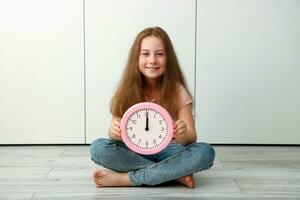 ein Mädchen Sitzung auf das Fußboden mit ein froh Lächeln hält ein Uhr Das sagt genau 12 Uhr foto