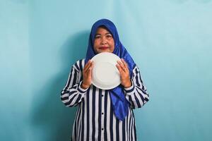 ein unzufrieden mittleren Alters asiatisch Frau im ein Blau Hijab und ein gestreift Hemd ist halten ein leeren Weiß Platte, Gefühl hungrig während ein Diät oder Fasten. sie ist isoliert auf ein Blau Hintergrund foto