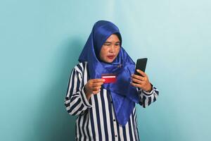 lächelnd mittleren Alters asiatisch Frau im Blau Hijab und gestreift Hemd halten Handy, Mobiltelefon Telefon und Anerkennung Karte, online Einkaufen isoliert Über Weiß Hintergrund. foto