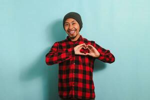 lächelnd jung asiatisch Mann zeigt an ein Liebe Geste mit seine Hand während Stehen gegen ein Blau Hintergrund foto